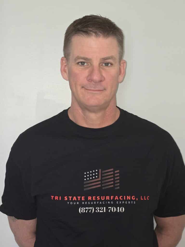 The image shows a person with short-cropped hair wearing a black T-shirt with an American flag and text, standing against a white wall.