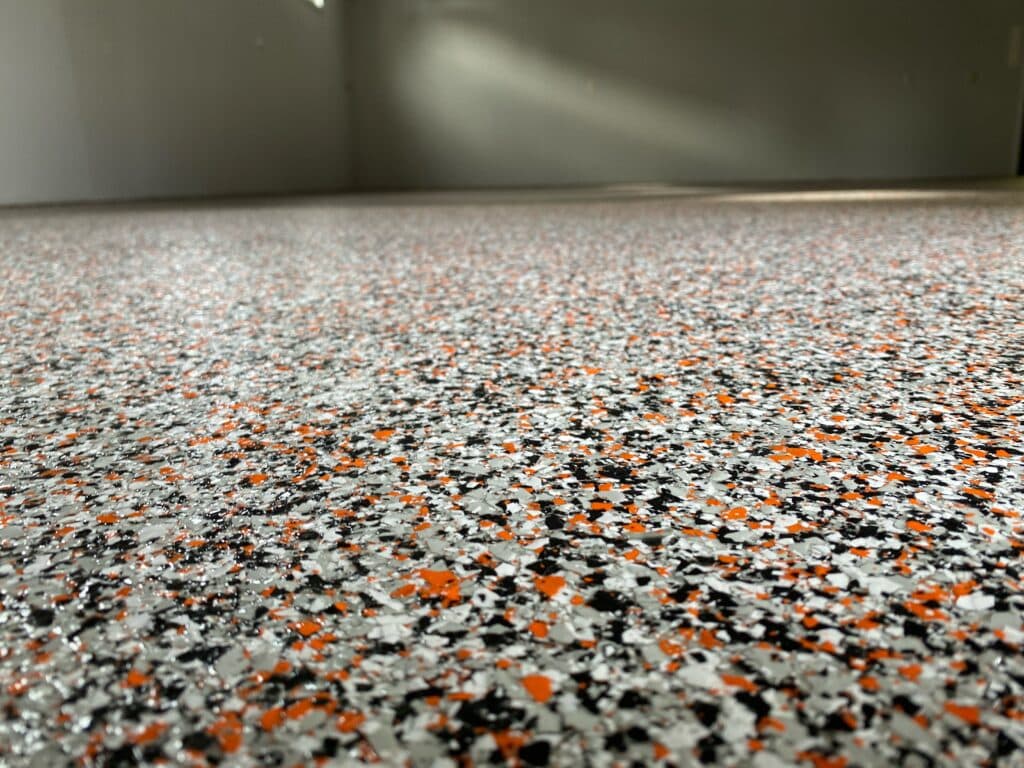The image shows a close-up of a speckled floor with a mix of orange, black, and white spots. Light casts a soft beam across the surface.